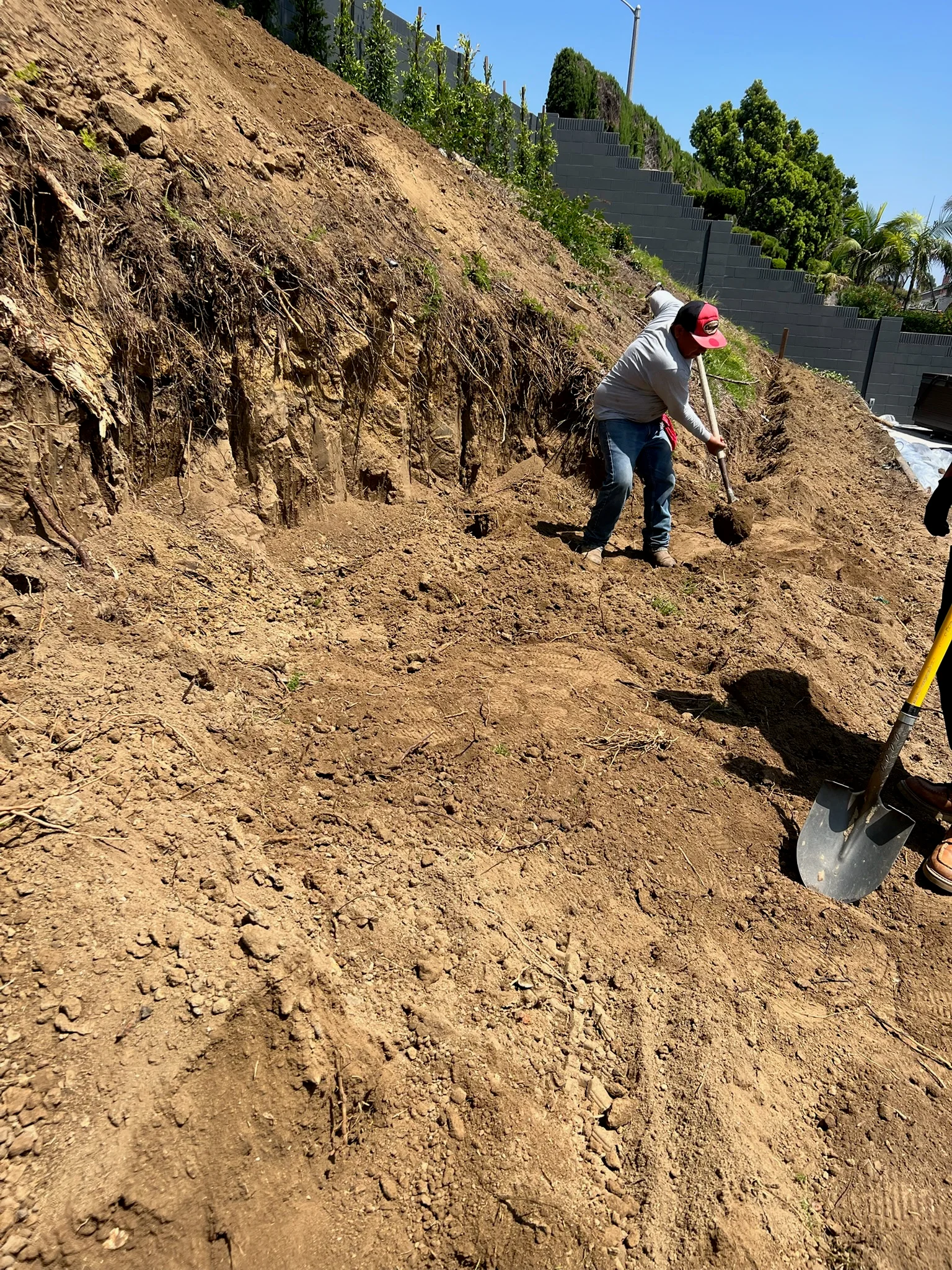Worker grading hillside soil with shovel; tiered retaining wall visible in background