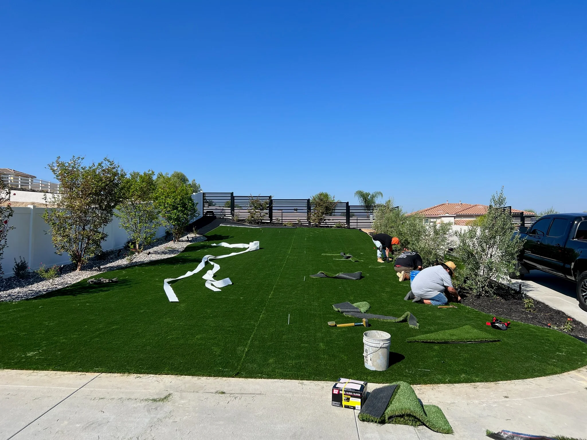 Synthetic turf installation in progress with black fence, olive trees, and landscape design
