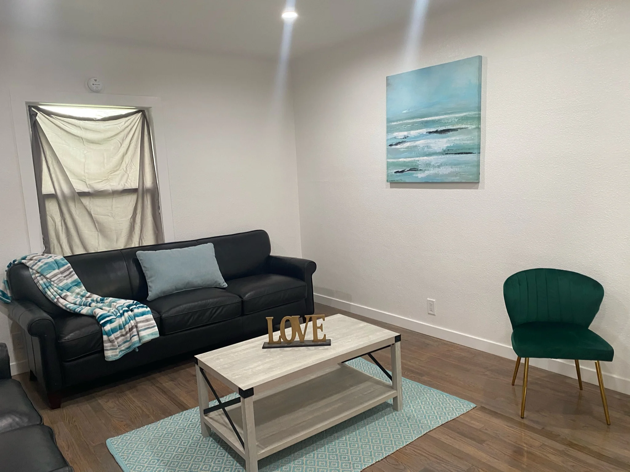 Renovated living room with hardwood floors, black sofa, light wood table, and coastal artwork