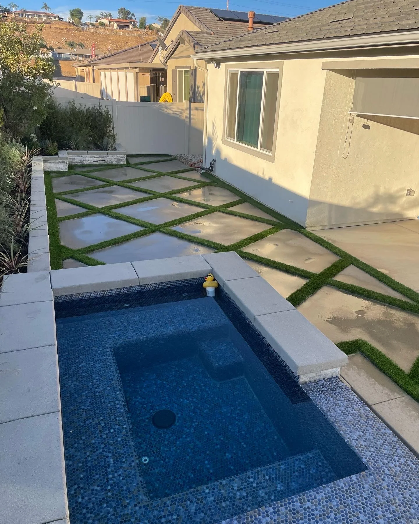 Modern backyard with blue tile pool, concrete patio with grass strips, and white fence