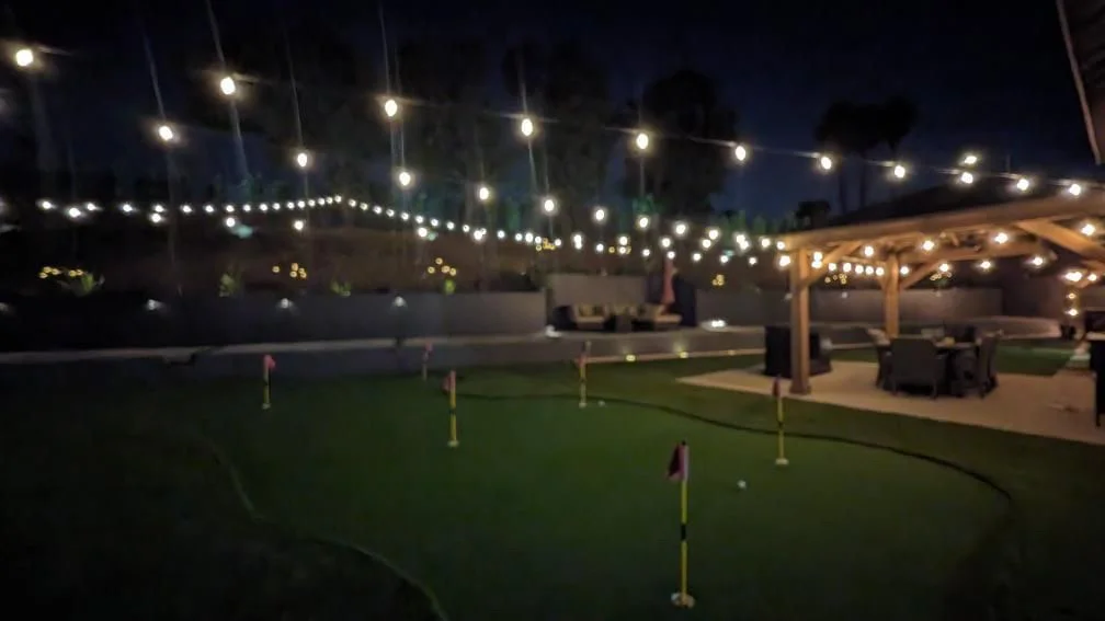 Nighttime backyard with illuminated putting green, string lights, and wooden pergola
