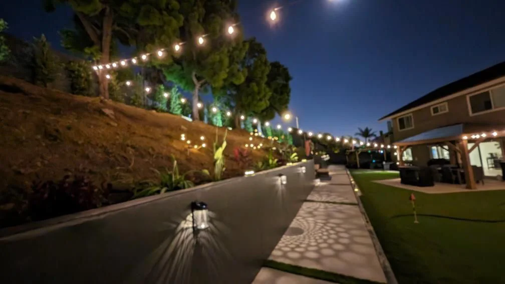 Illuminated backyard patio with concrete walkway, landscape lighting, and string lights at dusk