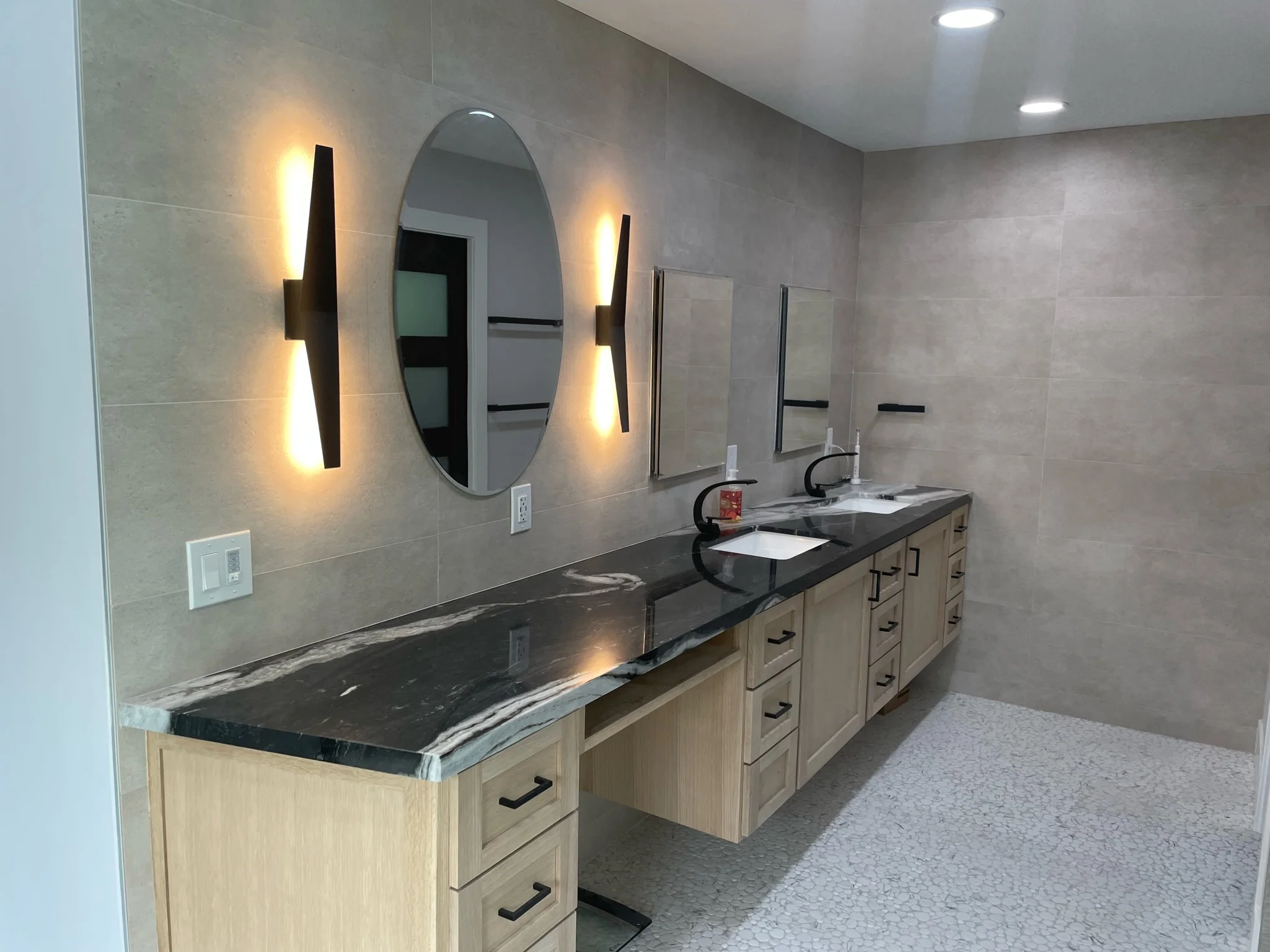 Modern master bathroom with dual vanities, black countertops, and gray tile walls