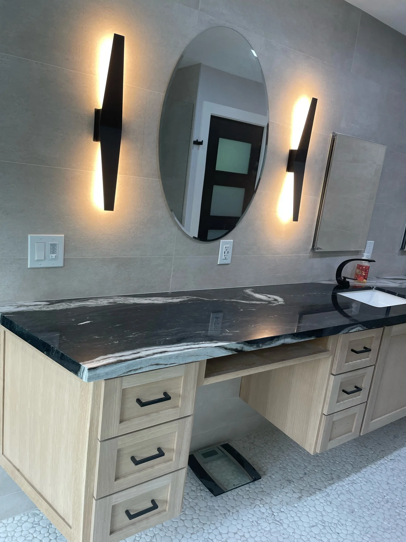 Modern bathroom with light wood vanity, dark marble countertop, oval mirror, and black wall sconces