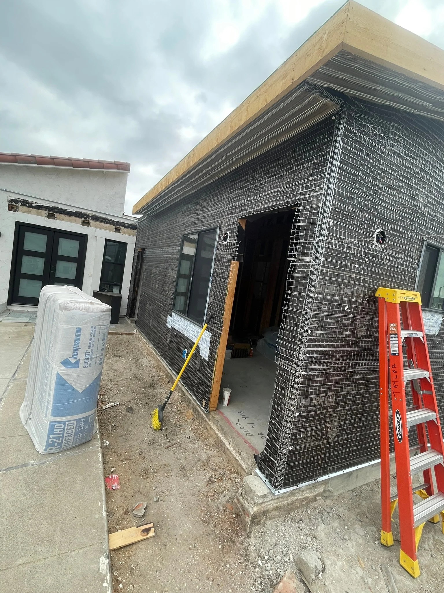 Modern home addition with black mesh exterior facade and wood-trimmed cantilevered roof under construction
